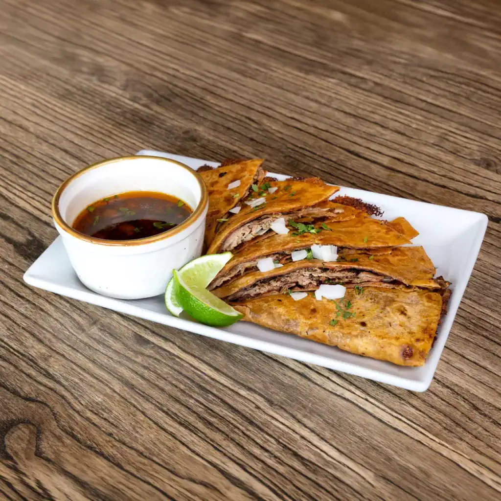 Grilled beef tacos with lime and dipping sauce on a white plate, wooden table background.